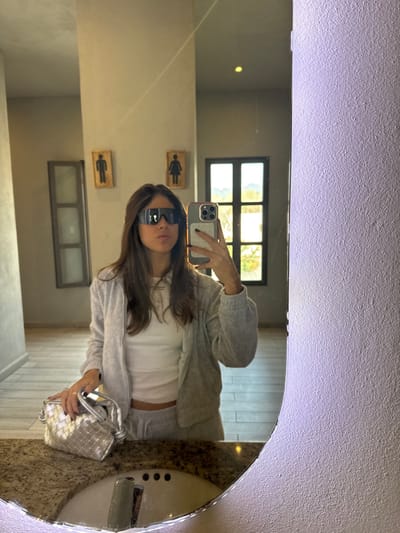 A woman in a white tank top and gray sweat suit takes a mirror selfie in a bathroom. She wears large sunglasses and holds a silver woven handbag. The bathroom features light-colored walls, a window, and gender symbol plaques. The mirror is round and illuminated. The image is a reflection.