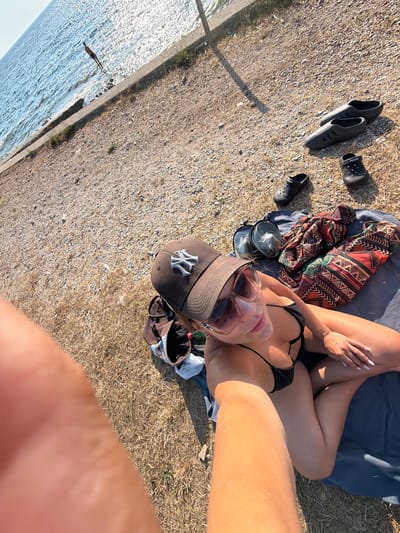 A woman is sunbathing on a blue towel spread on the ground near the coast in Buljarica, Montenegro. She is wearing a black bikini and sunglasses, with a brown baseball cap on her head. The ground is covered with pebbles and dry grass. Beside her, on the towel, are a pair of black and red patterned fabric and a diving mask. A person is visible standing in the water in the distance. Several pairs of black sandals are scattered on the pebbly ground to the right of the woman. The sun is casting long shadows.
