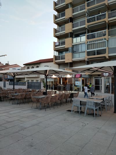 This media file captures an outdoor seating area of a restaurant in Canet-en-Roussillon, France, likely in the late afternoon or early evening given the soft lighting. The scene is dominated by numerous tables and chairs, both wicker and modern plastic, set up on a paved stone patio. Several large, modern cantilever umbrellas with integrated lighting provide shade over the tables, suggesting a comfortable dining experience.

In the background, a multi-story building with balconies and beige facade elements rises prominently. Adjacent to the building is a lower structure with a red-tiled roof, possibly another part of the restaurant or a neighboring establishment. The restaurant entrance is visible, with a sign that reads "restaurant LES FRAYEttes" and another smaller sign for "Artisan du GOUT." Further to the right, another entrance is marked with a sign that includes "LA CRÊPE" and "CRÊPES - GAL."

A few people are visible, primarily walking or standing near the restaurant entrances. Two individuals are walking away from the camera towards the right side of the frame, one wearing a striped shirt and the other a white top. Another person is visible inside the restaurant. A blackboard with handwritten text stands near one of the entrances. The overall atmosphere appears relaxed and inviting, hinting at a pleasant evening meal. The sky is a pale blue, indicating clear or partly cloudy weather.