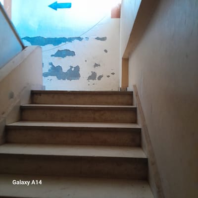 The image depicts a concrete staircase leading upwards, with several worn steps visible. The walls are plastered and show signs of age and damage, with patches of peeling paint, revealing a blue layer underneath in some areas. A blue arrow pointing to the left is painted on the upper portion of the wall, suggesting a directional sign. A wooden railing is partially visible along the right side of the staircase. The overall environment appears to be an indoor common area or hallway. The bottom of the image displays the text "Galaxy A14". The location is stated as Deir el-Balah, Palestinian Territories.