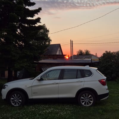 The image captures a white BMW SUV parked on a grassy area next to a building. The scene is set during sunset, as indicated by the warm orange and pink hues in the sky and the silhouetted trees. The car is positioned at an angle, with its side and rear visible. The building in the background appears to be a house, possibly in a rural or suburban setting, with a grey roof and some visible windows. Power lines are also present in the sky, stretching across the frame. The overall mood is tranquil and serene, fitting for a sunset. There is no visible text in the image.