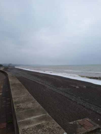 The image depicts a view of the Black Sea coast in Kobuleti, Georgia, on an overcast day. The foreground features a concrete embankment or seawall, with cobblestone paving visible to its left. Beyond the embankment is a dark, pebbly beach extending towards the sea. Gentle waves with white foam are breaking on the shore. In the distance, the sea merges with a hazy, grey sky. The overall impression is one of a tranquil, unpopulated coastal scene.