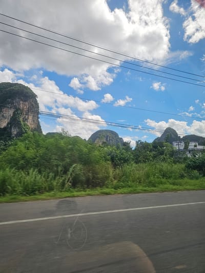 The image captures a scenic view from a moving vehicle, likely a bus or car, on a sunny day. The perspective is from the passenger side, looking out. The foreground features a paved road with a white line marking the lane. To the left of the road, lush green vegetation lines the roadside, interspersed with tall, rocky karst mountains characteristic of the Ao Luek region in Thailand. These mountains are covered in greenery, with some bare rock faces visible. In the distance, several buildings, possibly houses or hotels, are nestled among the trees on one of the larger karst formations. Overhead, a bright blue sky is dotted with fluffy white clouds. Several power lines are strung across the frame, adding a man-made element to the natural landscape. The overall impression is one of travel through a tropical, mountainous region on a clear day. The image appears to be taken during daylight hours. There is no visible text in the image.