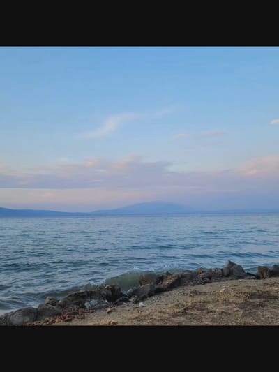 The image depicts a tranquil body of water, likely a lake, under a pale blue sky with wispy pink and white clouds. In the distance, a hazy mountain range is visible on the horizon. The foreground features a rocky shoreline with dry grass and scattered debris. The water is calm with gentle waves lapping against the shore. The overall scene suggests a serene natural environment. The image does not contain any people or specific urban elements that would indicate a location within Bielawska Wieś, Poland.
