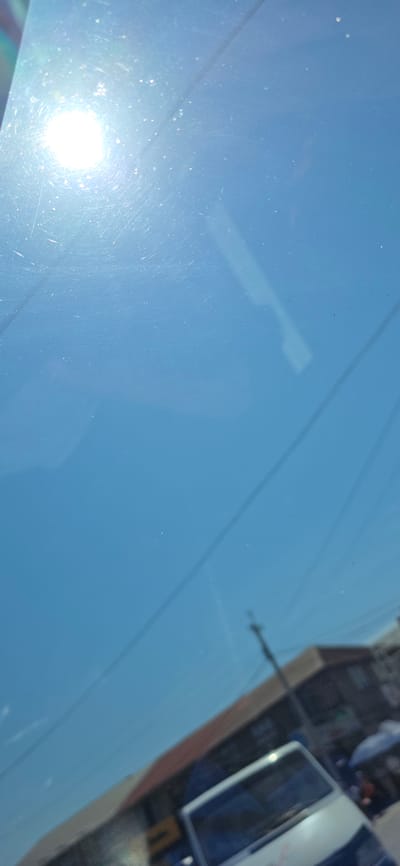 The image is a vertical, outdoor photograph looking upwards at a bright, sunlit sky, likely through a window. The sun is prominently visible in the upper left quadrant, casting a strong glare and creating lens flares and small circular light spots. The sky is a clear, bright blue, with subtle reflections and scratches visible on the surface through which the image is taken. In the lower portion of the image, a blurred, diagonal line suggests the top edge of a building or structure. Further down and slightly to the right, the blurred shapes of buildings with reddish-brown roofs and awnings are discernible. A white van, also out of focus, is positioned in the lower right quadrant, with its windshield reflecting the bright sky. The presence of a utility pole is also visible. The overall impression is a snapshot taken during the day in an urban environment, possibly from inside a vehicle or building, with the sky and sun being the dominant elements. Given the specific request for the city of Karakol, Kyrgyzstan, it is likely this scene depicts a street view in that location, characterized by typical urban architecture and daytime conditions.