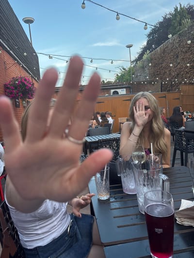 Outdoor scene with a group of people at tables. A young woman covers her face with her hand while seated at a table with multiple glasses. The foreground is dominated by an outstretched hand with a ring. A denim skirt and white top are visible on the right. String lights hang overhead against a backdrop of a building and trees.