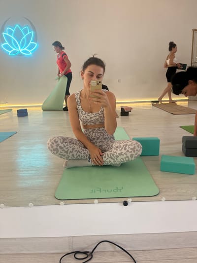 In a brightly lit yoga studio, a woman in a leopard-print athleisure set sits cross-legged on a mint green yoga mat. She holds a smartphone in her right hand, taking a selfie. Her gaze is directed towards the phone's screen. She wears a silver cross necklace and a ring on her left hand. Several other women are visible in the background, engaged in yoga activities. One woman in a pink shirt is unrolling a mat, while another in a white top and black shorts walks by. A neon lotus flower sign glows on the white wall. The floor is made of light-colored wood planks, and various yoga blocks are scattered around the mats. The scene is a reflection in a large mirror that runs along the bottom of the room. The location is the city of Aksu, Kazakhstan.