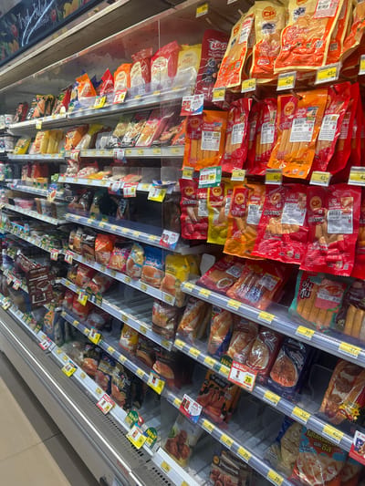 The image shows an aisle of a convenience store, likely a 7-Eleven, in Ban Kong It, Thailand, as indicated by the product branding and Thai text. The shelves are stocked with a variety of pre-packaged food items, predominantly sausages, hot dogs, and other processed meat products. The products are arranged in colorful packaging, with prominent branding such as "CP" and various Thai text. Many items have price tags displayed, with prices ranging from 29 to 45 Baht. The lighting is fluorescent and typical of an indoor retail environment. There are no people visible in the frame. The overall impression is of a well-stocked food section in a typical Thai convenience store.