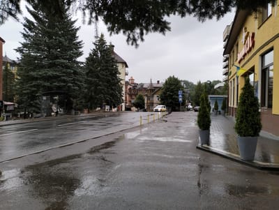 The image depicts a street scene in Truskavets, Ukraine, on a cloudy day after rain, as evidenced by the wet asphalt and puddles. On the right side of the frame, a yellow building with red signage and Ukrainian flags displayed is visible. In front of the building, two large grey planters contain tall, conical evergreen trees. A paved sidewalk runs alongside the building.

To the left of the building, across the wet street, are several other buildings with varied architectural styles, including some with ornate balconies and towers. A bus stop shelter with a person sitting inside is located further down the street on the left. Tall evergreen trees line the street, some obscuring parts of the buildings. Traffic signs, including a blue sign with white symbols indicating pedestrian crossing and direction, are present along the road. A white car is parked or stopped on the right side of the road. Yellow bollards are spaced along the edge of the road or sidewalk. The sky is overcast and grey, contributing to the muted light of the scene.
