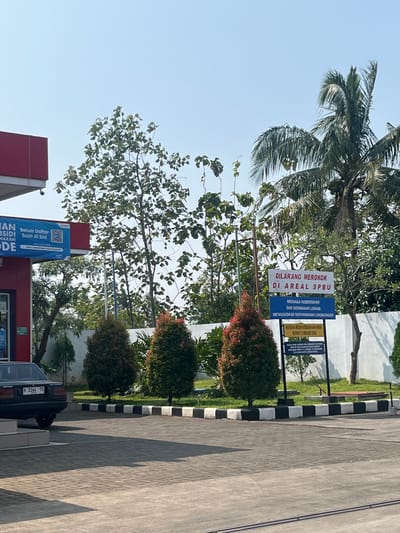 This is a wide shot of an outdoor scene on a sunny day, likely in Meteseh, Indonesia, given the signage. The foreground is paved with light gray interlocking bricks, casting long shadows from the right. A dark-colored sedan is parked on the left.

In the middle ground, a row of three conical evergreen trees with reddish-brown tips lines a black and white checkered curb. Behind these trees is a white wall.

On the right side of the image, a tall palm tree with lush fronds dominates the upper portion. To its left, a series of three signs on metal posts are visible. The top sign, in red and white, reads "DILARANG MEROKOK DI AREAL SPBU" (No Smoking in the SPBU Area). Below it, a blue sign states "MENJAGA KEBERSIHAN DAN KEINDAHAN LOKASI MEWUJUDKAN KENYAMANAN LINGKUNGAN" (Maintaining Cleanliness and Beauty of the Location Creates Environmental Comfort). The lowest sign, in yellow and black, advises "MATIKAN MESIN KENDARAAN ANDA SEWAKTU MENGISI BBM" (Turn off Your Vehicle Engine While Refueling). Further text is present on the yellow sign but is not legible.

A red and white building, possibly a gas station (SPBU), is partially visible on the far left, featuring a blue banner with Indonesian text and a QR code. Several trees with green foliage are scattered throughout the scene, with thinner, vine-like plants also present. The sky is clear and light blue.