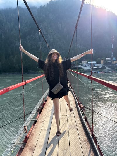 A woman with long, curly brown hair, wearing a black t-shirt, black skirt, a straw hat, and brown sandals, stands on a suspension bridge with red railings and a wooden walkway. Her arms are outstretched, and her right leg is lifted. A beige Gucci crossbody bag hangs from her left shoulder. The bridge spans a wide river with glistening water. In the background, lush green mountains rise, partially obscured by mist. A few buildings and a cell tower are visible on the far bank of the river. The sun is setting, casting a warm glow and lens flare across the scene. The location is the suspension bridge over the Katun River in the village of Barangol, Russia.