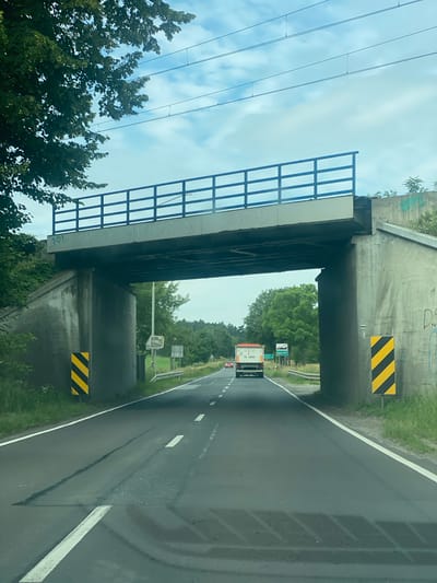 This image depicts a view from a vehicle driving on a two-lane road under a concrete overpass. The overpass has a blue metal railing on top and is supported by two large concrete pillars, one on each side of the road. The pillars have diagonal black and yellow striped warning signs attached. Graffiti is visible on the underside of the overpass and on the concrete supports.

In the distance, on the road, a white truck with orange accents is visible, followed by a red car. Traffic signs are present on the right side of the road, one of which indicates "Miłosław" with an arrow pointing to the right, suggesting the location is near or in Miłosław, Poland. The surrounding environment is green with trees and foliage on either side of the road and beyond the overpass. The sky is cloudy with blue patches. The road itself shows signs of wear with patched areas and visible lane markings. Electrical wires are strung across the sky above the overpass.