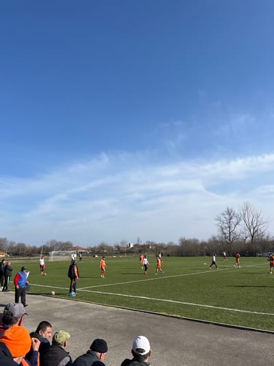 This image depicts an outdoor football match in progress on an artificial green turf pitch located in Kliment, Bulgaria. Approximately 10-12 players are visible, divided into two teams: one wearing orange kits and another in white shirts with maroon shorts or accents. Players are distributed across the field, with white lines marking the pitch boundaries.

In the foreground, a group of male spectators, some wearing hats, are seated or standing, observing the game from behind a concrete barrier. Near the sideline, a male individual in a blue vest and red shirt stands, appearing to be a coach or official, observing the play. A football rests on the pitch near the sidelines in the mid-ground.

The background features distant residential buildings, including a structure resembling a church tower, and numerous bare deciduous trees, suggesting late winter or early spring. A white goalpost is visible on the left side of the field. On the far side of the pitch, a sign with the word "ELIT" is discernible. The sky is predominantly clear blue with sparse, thin white clouds, indicating bright daylight conditions. Shadows are visible on the field and among the spectators, consistent with direct sunlight.