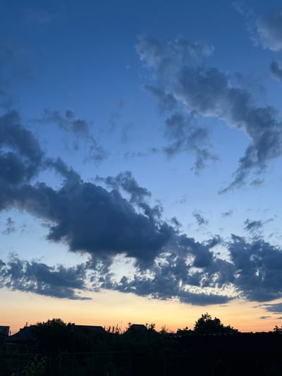 The image depicts a dusky sky over a residential area, likely in Slobozia, Moldova. The upper portion of the frame is dominated by a deep blue sky transitioning to lighter hues near the horizon, with scattered dark grey clouds creating a dramatic effect. The lower part of the image shows a silhouetted landscape of rooftops and dense foliage, suggesting a rural or suburban setting. The overall impression is one of twilight, with the last light of day casting a warm glow near the horizon. There are no visible people or specific actions taking place, making it a static environmental scene. The objects present are houses, trees, and a fence in the foreground, all rendered in dark silhouettes against the brighter sky.