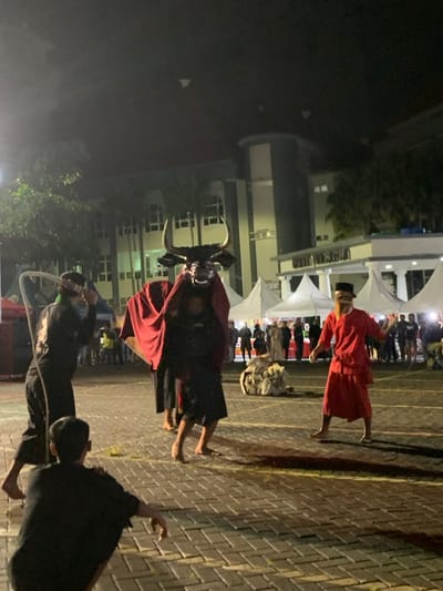 The image captures a performance in an outdoor area in Kecamatan Batu, Indonesia, located at latitude -7.867 and longitude 112.514. The performance appears to be a traditional Indonesian dance, possibly a re-enactment of a mythical event. A person dressed in black and red clothing is carrying a large black bull's head mask on their shoulders, with red fabric draped over the mask and down to the floor. The person is walking towards the right of the image. To the left of the dancer, a man dressed in black is holding a rope and watching the dance. Another man, wearing a black shirt and pants, is sitting on the ground watching the performance. In the background, a large building with a white awning is visible, with several white tents set up in front of it. Several people are standing around the tents, watching the performance. The stage is illuminated by artificial lighting, casting a warm glow on the performers and audience.