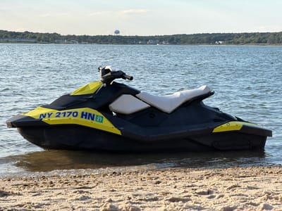 A yellow and black jet ski is partially submerged in shallow water at the edge of a sandy and pebbly beach. The jet ski has the registration number "NY 2170 HN" and a green sticker with the number "27" on its front left side. The water is calm with small ripples, and the sky is clear with a few scattered clouds. In the background, a tree-lined shore is visible with some houses and a water tower in the distance, suggesting a location in Hampton Bays, United States of America. The lighting indicates it is daytime, likely late afternoon due to the warm tones.
