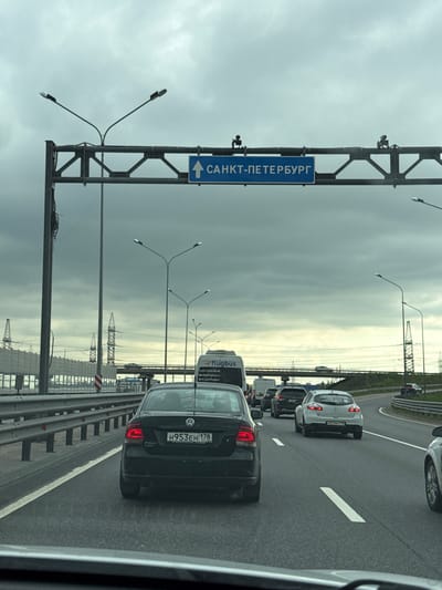 The image shows a traffic jam on a highway, likely a route leading to Saint Petersburg, Russia. A blue road sign indicating the direction to "Saint Petersburg" (САНКТ-ПЕТЕРБУРГ) is visible. Several cars are lined up, including a black Volkswagen Polo with the license plate "H953EH 178" and a white car with the license plate "M023BO 178." A Flubus bus is also in the lineup. Streetlights and power lines are on the side of the road. The sky is overcast. The presence of power lines and industrial structures in the background suggests an area on the outskirts of a city. It seems to be a highway on a bridge. The surroundings are urban. Korabsel'ki, Russia is not mentioned in the information or visible in the image.