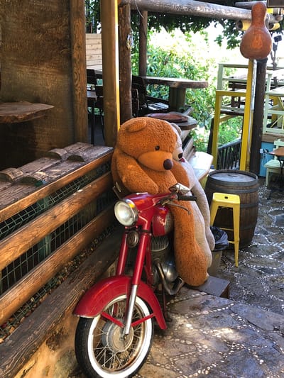 The image depicts an outdoor seating area with a rustic, bohemian aesthetic, likely located in Kaş, Turkey. In the foreground, a vintage red motorcycle is parked. Seated on the motorcycle are two large teddy bears, one brown and orange, the other a lighter tan. The brown teddy bear's arm is draped over the motorcycle's handlebars. Behind the motorcycle, a large wooden barrel stands next to a bright yellow stool and a black trash can. Wooden fencing and benches are visible to the left, with carved wooden details on the bench. In the background, an array of tables and chairs are set up amidst lush green foliage, suggesting an outdoor cafe or restaurant. Sunlight filters through the trees, casting dappled shadows on the stone-paved ground. A decorative gourd-shaped lantern hangs from an overhead wooden structure, adding to the unique ambiance.