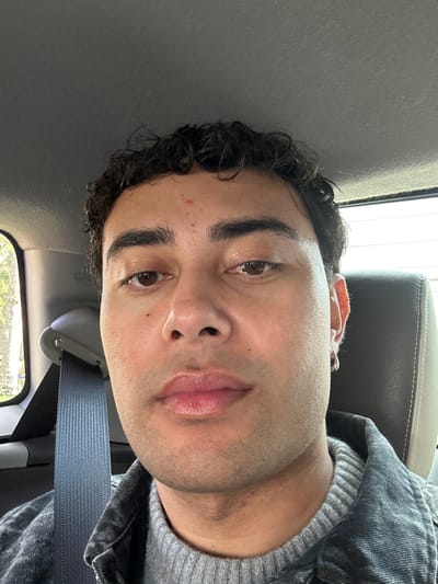 An individual is depicted in a close-up portrait, seated inside a vehicle in what is identified as Neuilly-Plaisance, France. The subject is a male with olive skin, dark curly hair, and dark brown eyes. A small reddish blemish is present on his forehead. He wears a small silver hoop earring in his left ear. His attire consists of a grey ribbed knit sweater under a dark, possibly denim or canvas, jacket. A light blue seatbelt strap extends diagonally across his left shoulder and chest. The vehicle's interior features a grey headliner and a padded light-colored headrest visible to the right of the subject's head. Through the vehicle's window on the left, blurred green foliage and ambient light are discernible. The individual maintains a neutral facial expression, looking directly towards the camera. No other persons or specific actions are visible.