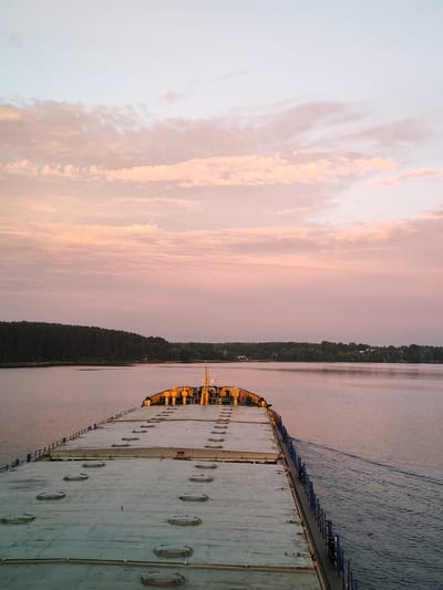 The image captures a wide-angle view of a barge moving through a calm body of water, likely a river or lake, at dusk. The sky is painted with soft hues of pink and pale blue, indicative of sunset or sunrise, with scattered, wispy clouds. The barge, seen from a slightly elevated perspective on its deck, is the primary subject in the foreground. Its deck is a flat, weathered surface with numerous circular hatches, suggesting it is a cargo barge. The stern of the barge, adorned with yellow structures and a mast, is visible further down the deck.
The water is a muted, reflective surface, with faint ripples trailing behind the barge, indicating its movement. On the horizon, a densely forested treeline stretches across the landscape, forming a dark green band against the softer sky. The trees appear to be on the shore of the body of water. Subtle hints of structures, possibly buildings or other human presence, are scattered among the trees, suggesting a rural or semi-rural settlement. The overall atmosphere is serene and peaceful, dominated by the soft light and the gentle movement of the barge. The location is identified as Терехово, Russia, suggesting this scene is situated within or near this Russian locality.