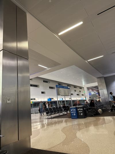 This indoor shot shows the inside of an airport with people waiting in the terminal. The ceiling is high and white with rows of recessed lighting. Below it, a stainless steel-looking structure is featured at the bottom of the image. Multiple rows of seating face towards the windows. People are sitting and waiting. Several signs are visible. The location is likely an airport in Fort Lauderdale, United States. The overall composition is angled, giving an unusual perspective of the interior space.