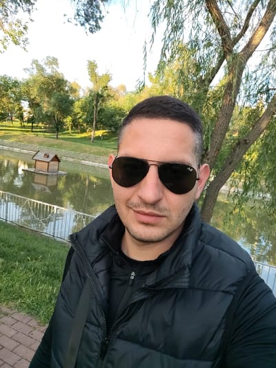 A man wearing dark sunglasses and a black puffy vest stands in front of a pond or small lake in a park. The pond has a small, decorative birdhouse in the middle. A metal fence borders the pond. Trees surround the pond. The location is Taraclia, Moldova.