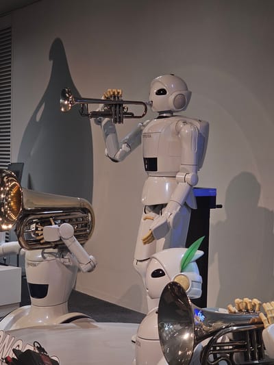 In a museum setting in Nagoya, Japan, three humanoid robots are depicted with musical instruments. The robot in the foreground left holds a tuba, its robotic arms supporting the instrument. A second robot, positioned centrally, plays a trumpet, its hands on the valves and its mouth near the mouthpiece. A third robot, partially visible in the background right, also appears to be holding a musical instrument. The robots are white and sleek, with visible joints and articulated hands. They are positioned on a floor that appears to be carpeted, with a plain gray wall as the backdrop. The lighting casts shadows on the wall, adding depth to the scene. The robots are branded with the "TOYOTA" logo.