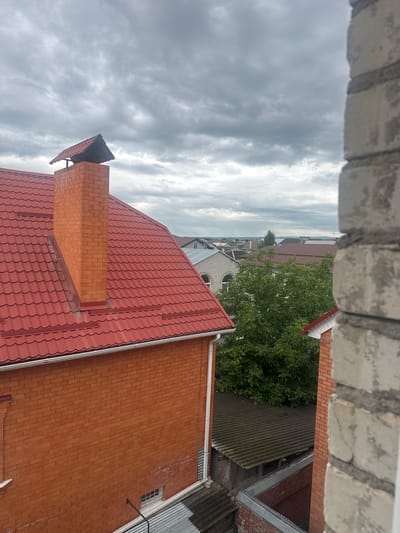 An aerial view of a residential area in the city of Svobody, Russia, is captured under a cloudy sky. In the foreground, the bright red, corrugated metal roof of a building is prominent, with a red brick chimney rising from it. The chimney is topped with a dark, angled cap. To the right, a partial view of a light-colored brick wall, likely part of a window frame or adjacent building, occupies the edge of the frame. Beyond the immediate structures, a dense green tree provides a vibrant contrast to the muted tones of the sky and roofs. In the middle ground and background, numerous other houses with red and grey roofs, along with some glimpses of a body of water and distant landscape, are visible. The overall impression is of a suburban or semi-rural setting on an overcast day. There are no people visible in the image.
