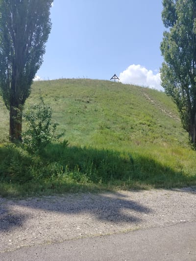 The image depicts an outdoor scene dominated by a grassy hill under a bright blue sky with scattered white clouds. Two tall trees, one on the left and one on the right, frame the hill. The tree on the left is closer to the foreground and has a thick trunk and dense foliage. The tree on the right is partially visible at the edge of the frame. A small bush or sapling is situated near the base of the left tree. The hill itself is covered in green grass, with a visible path winding up its slope. At the crest of the hill, a small wooden cross-like structure is present. In the foreground, a gravel path transitions to an asphalt road. The overall impression is of a rural or park-like setting. The image does not contain any people or visible signs of recent activity. The location in Balatina, Moldova, is not directly indicated by any visible landmarks or text within the image itself.