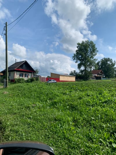 The image shows a rural scene in the city of Barsai, Russia, on a sunny day with scattered clouds. The foreground is dominated by a lush, green meadow. A utility pole with wires stands on the left side of the image. Behind the meadow, there is a small, light-colored house with a red roof and trim, and several red-striped fences. A silver car is parked near the fence. Further in the background, there are several trees and other buildings, including a long, light-colored structure with a red roof. The overall impression is one of a tranquil, semi-rural environment.