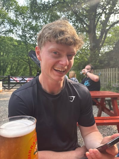 The image shows a man wearing a black shirt with a white "GYM" logo, sitting at a red picnic table in a beer garden at the "The Royal Oak" pub, in Hayfield, United Kingdom. The man is holding a beer glass and looking at a smartphone. He is smiling and has short, light blonde hair. He is seated at a table outside the pub, with a man in a blue shirt sitting in the background. The setting is outdoors, with trees visible in the background. The scene is relaxed and social, with the men enjoying a drink together. The man in the foreground appears to be enjoying his time. The pub is located at 53.377 degrees latitude and -1.935 degrees longitude.