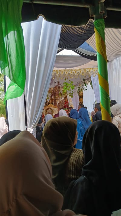 Captured from a low angle inside what appears to be a tent-like structure, the scene focuses on an event occurring within, possibly a wedding. Observers are gathered around. The foreground is dominated by the backs of individuals, their clothing ranging in colors. A central backdrop features a golden-trimmed curtain, and green foliage is placed in front. Hints of architecture and open space are visible through the tent's structure, suggesting an outdoor setting. The location is Pulo, Indonesia.