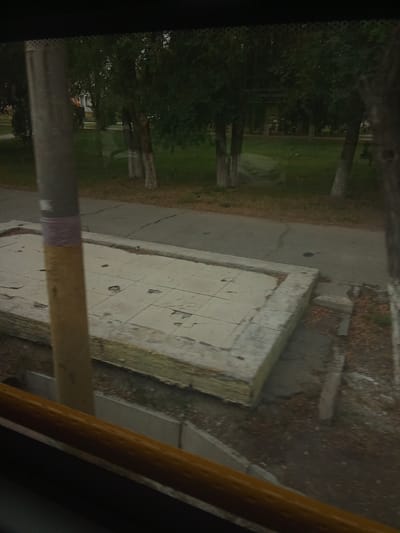 The image depicts an outdoor scene, likely in the city of Taraz, Kazakhstan, seen through a window. In the foreground, a brown-colored handrail is visible, suggesting the viewpoint is from inside a vehicle or building. To the left, a vertical gray pole with yellow and purple bands stands prominently. This pole is positioned in front of a raised, rectangular concrete structure covered with square tiles. The structure appears somewhat damaged or unfinished, with visible layers of insulation beneath the tiles.
Beyond this structure, a paved road with cracks is present. Behind the road, a grassy area is visible, dotted with several trees. The trees have light-colored bark, and their foliage is dark green. The overall environment suggests a park or a tree-lined street. Reflections on the window obscure some details of the background, but hints of buildings and further trees can be discerned. The lighting suggests it is daytime, possibly overcast.