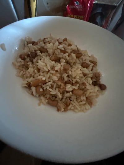 The scene, located in the city of Egbe, Nigeria, displays a white, circular bowl centrally filled with a cooked meal. The meal consists of light-colored rice mixed with numerous small, light brown beans. The bowl itself is plain white with a subtly curved rim. The background elements are out of focus. In the upper left background, a yellow and brown patterned object is partially visible. In the upper right background, a red flexible package or pouch with the visible text "Good Earth" and "HOT PEPPE" is present. No people are depicted. The lighting indicates an indoor setting.