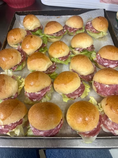 The image displays a close-up, eye-level view of a metal baking tray filled with freshly made mini sandwiches. The sandwiches are arranged in multiple rows and consist of golden-brown, glossy buns split open to reveal layers of fresh green lettuce and thinly sliced salami. Some of the salami slices have visible white fat marbling. The buns appear soft and slightly puffed, suggesting they are freshly baked. The baking tray itself is metallic and shows some minor signs of use. The background is out of focus but hints at a kitchen environment with hints of red and white kitchenware. The lighting is bright and even, illuminating the food items clearly. The scene is set in a commercial or home kitchen, likely in Italy given the prompt's request to include the location of Vallo della Lucania, Italy, though no specific landmarks are visible to confirm this. The photograph is taken from a slightly overhead angle, looking down onto the tray of sandwiches.