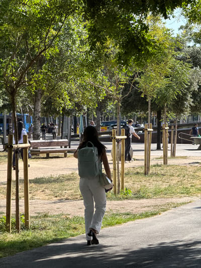A woman walks away from the camera, seen from behind, wearing white pants and a light blue backpack. She is on a paved path with shadows from trees overhead. To her left, a park-like area with dry grass and scattered trees is visible, with wooden posts lining a pathway. In the background, several people are walking and standing, and a bus is parked on a street. The scene appears to be an urban park or promenade in L'Hospitalet de Llobregat, Spain, with a sunny day and mature trees providing shade.