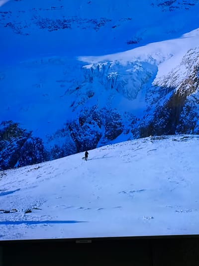 The image shows a person walking across a vast, snow-covered landscape with steep, icy mountains in the background. The sky is a deep blue, and the overall scene is one of harsh, frozen beauty. There is a distinct shadow cast by the person, indicating a bright light source, likely the sun, although it is not directly visible. The snow appears uneven, with footprints clearly visible in the foreground, suggesting a recent passage. The mountains are rugged and jagged, covered in snow and ice formations. There are no buildings or urban elements visible, and the setting appears to be a remote, mountainous region. The image does not depict any specific location in the city of Tewkesbury, United Kingdom, as Tewkesbury is a historic market town located in Gloucestershire, England, and is characterized by its timber-framed buildings and its position on the River Avon, not by a snowy mountainous terrain.