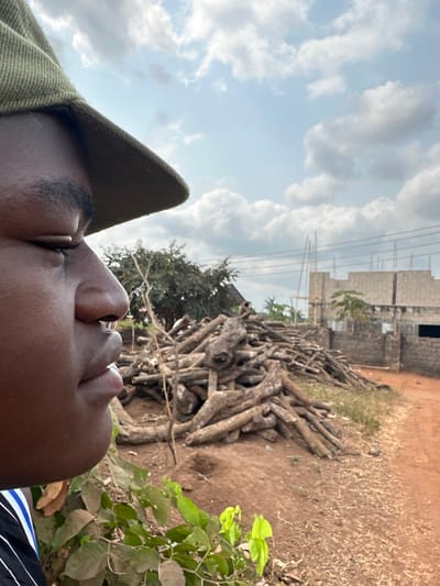 The image depicts an outdoor scene in Agbogugu, Nigeria. In the left foreground, the left profile of an individual with dark skin is visible, wearing a green cap and a striped shirt. The individual's eyes are partially closed. Green foliage is present in the lower left foreground. The midground features a large, irregular pile of cut wooden logs or tree branches resting on reddish-brown earth. To the right, an unpaved reddish-brown dirt path is visible. In the background, a building under construction, composed of concrete blocks, is present with rebar extending upwards. Overhead, power lines span horizontally across the upper right portion of the frame. The sky is partially cloudy. Sparse trees and brush are scattered throughout the background. No specific actions or interactions between individuals are depicted.