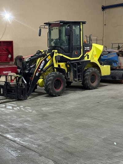 A yellow and black Digger wheel loader is located in an indoor area, possibly a warehouse or storage facility, in Shkotovo, Russia. The loader is partially facing the viewer, showcasing its cab, wheels, and loading arms. In the background, there is additional equipment, including a blue vehicle and red container. The floor is concrete.