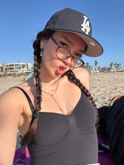 A female subject is depicted in a close-up selfie on a sandy beach in Manhattan Beach, United States. She wears a black baseball cap with a white "LA" logo, tortoise-shell framed eyeglasses, and a black camisole. Her dark hair is styled in two braids that frame her face. Visible accessories include a silver-colored hoop earring with a clear square drop on her left ear and multiple layered necklaces, one featuring a cross pendant. The subject is actively posing with a playful expression, characterized by pursed lips and a slightly extended tongue. The scene is illuminated by direct sunlight. In the background, a wide expanse of sand extends towards the ocean, under a clear blue sky. Distant low-rise buildings, palm trees, and a multi-story modern building are visible along the coastline. Other individuals are discernible further down the beach. A purple and white striped towel is partially visible to the lower left of the subject, and a dark bag or object is partially visible to the lower right.