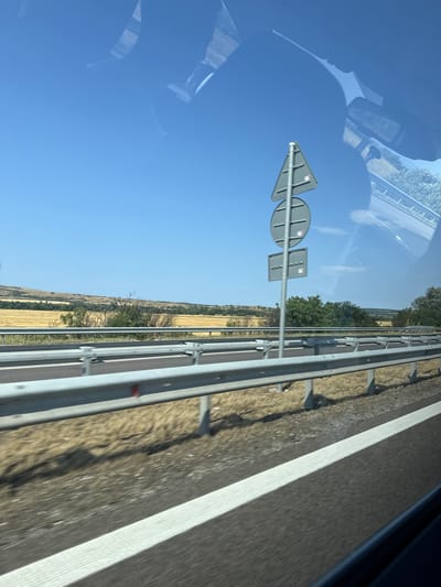 A roadside view from a moving vehicle in Kalugerovo, Bulgaria, shows a clear blue sky above a landscape of dry, golden fields and scattered trees. A metal guardrail runs horizontally across the foreground, separating the road from the field. A triangular traffic sign with a circular sign below it stands near the guardrail. The road surface, marked with white lines, is visible in the lower portion of the image. Reflections of the vehicle's interior, including parts of the dashboard and possibly the sky, are visible on the window through which the photograph was taken. The scene suggests a rural, possibly agricultural area under a bright, sunny sky.