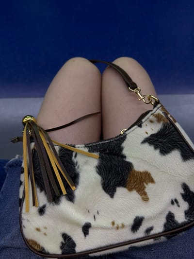 A close-up, low-angle shot shows a person's legs and a handbag. The person is wearing denim shorts. The handbag is made of faux cowhide and has dark brown and yellow fringe. A dark brown strap with a gold clasp is attached to the bag. The background is a solid blue color. The image does not contain information about the location in Dongducheon-si, South Korea.