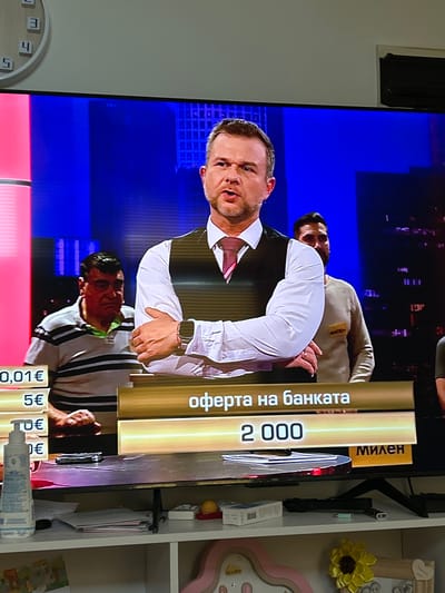 The image shows a television screen displaying a game show or similar program, captured from a slightly angled perspective and rotated 90 degrees clockwise.
On the television screen, three men are visible. The most prominent figure, centered, is a man with short brown hair and a beard, wearing a white collared shirt and a dark vest, looking intensely forward. Above him, another man in a light-colored sweatshirt with a "МИЛЕН" (Milen) name tag is partially seen. Below the central figure, a third man in a black and white striped shirt is visible from the chest up. The background on the TV is a bright blue, suggesting a studio setting.
Prominently displayed on the right side of the screen are two golden vertical bars. The left bar reads "оферта на банката" (offer from the bank) in Cyrillic, and the right bar shows the number "2 000". Along the bottom edge of the screen, several smaller vertical bars display monetary values in Euros: "0,01€", "5€", "10€", and another partial "€" amount.
The television itself is surrounded by a home environment. To the left, a plain light-colored wall is visible. To the right, a white shelf unit holds several decorative items, including a yellow fabric flower, a pink heart-shaped frame with the date "06.11.20258" inscribed, and a white bottle, possibly a lotion, with "Chantsue" visible on its label. A wall clock, partially visible on the bottom left wall, shows some Cyrillic letters like "LT" and "Ш". The visible Cyrillic text and Euro currency are consistent with the location context of Kavarna, Bulgaria.