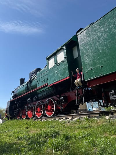 The image captures a green and red steam locomotive, likely a preserved exhibit, situated on train tracks amidst green grass. The sky is clear and blue. A young boy in a red shirt, light-colored pants, and a white hat is climbing on the train. The photograph was taken in Balezino, Russia.