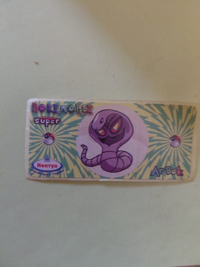 This is a close-up, overhead shot of a Pokemon card, lying on a light green, textured surface. The card has a yellow background with radiating teal lines, resembling stylized lightning bolts.
The main subject on the card is a purple Pokemon, identified as Arbok by text at the top right of the card. The Pokemon is depicted coiled in a pinkish circle. At the top of the card, between the Arbok name and the Pokemon image, is a small Pokemon ball. Another Pokemon ball is visible at the bottom center of the card, just below the image.
To the left of the Pokemon image, vertically oriented text reads "No.NMON.super". Below this text, and to the right of the bottom Pokemon ball, is a blue oval with red text that appears to read "Нептун". There are also some smaller, illegible words in white text within the blue oval.
The card itself appears to be slightly worn and creased, particularly at the bottom edge. The lighting suggests it is daytime, but the setting is indistinct beyond the surface the card is placed upon. There are no people visible.