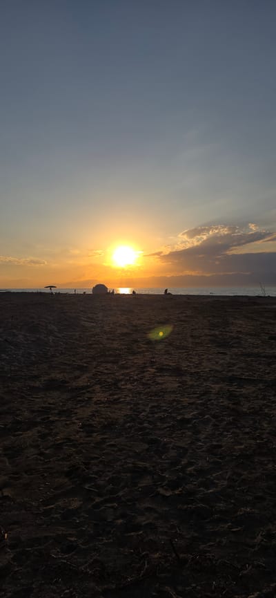 The image depicts a serene sunset over a beach. The sun, a bright orb, is positioned just above the horizon, casting a warm, golden glow across the sky and reflecting on the water. The sky transitions from a deep blue at the top to hues of orange and yellow near the horizon. Scattered clouds are visible, catching the sunlight.
In the foreground, a sandy beach stretches across the frame. The sand appears dark and textured, with visible footprints and debris. In the middle ground, several figures are present on the beach, some sitting or standing, silhouetted against the bright sunset. An umbrella is visible to the left of the frame. A large, rounded object, possibly a rock or a large object, is situated on the beach near the water's edge. The water itself appears calm, with gentle waves lapping the shore. The distant horizon is marked by a range of mountains, silhouetted against the colorful sky. The overall scene suggests a peaceful end to the day, with people enjoying the natural beauty of the beach at sunset. The location provided, Boz Beshik, Kyrgyzstan, indicates a setting in Central Asia, known for its mountainous terrain and natural landscapes.