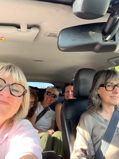 Five adult women are in a car. The woman in the front is blonde and wearing glasses, partially obscuring her face. In the second row, a woman with short white hair and sunglasses is smiling. Above her, partially obscured, is another woman. The woman in the third row is wearing glasses. All passengers are wearing seatbelts. The car's interior is visible, including the rearview mirror and sun visor. The location is Montélimar, France.