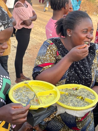 A group of people are gathered, seemingly enjoying a meal outdoors. A woman in a patterned dress is laughing, possibly eating. Two yellow divided plates filled with a green stew-like dish sit beside her. Someone is eating from one of the plates with a spoon. Another woman wears a pink shirt and black leggings. The ground is light-colored, possibly dirt or sand. A black purse is also visible.