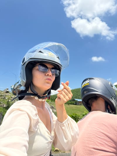 Two individuals, presumably on a motorbike, are captured in a selfie taken outdoors under a bright blue sky with scattered white clouds. The person in the foreground, wearing a grey helmet with a clear visor and sunglasses, is looking at the camera with their lips pursed and making a gesture with their hand that signifies "I love you" in Korean culture. They are also wearing a light-colored top and a seashell necklace. Behind them, partially visible, is another person wearing a dark helmet and a pink shirt. The background features lush green hills and some buildings, indicative of a rural or semi-rural setting. The presence of the helmets suggests they are traveling by motorbike, a common mode of transportation in Vietnam. The overall scene appears to be a casual moment captured during an outing, likely in a scenic location such as Hàm Thuận Bắc, Vietnam, given the terrain and vegetation.