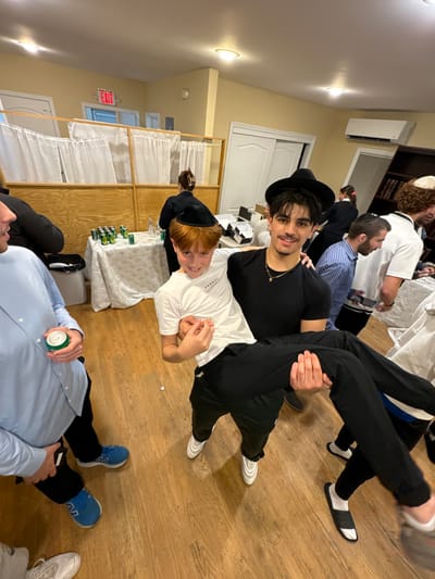 This image depicts an indoor social gathering at a facility in Bergenfield, United States. In the central foreground, a young adult male with dark hair, a dark fedora, and a subtle beard, wearing a black t-shirt and dark pants, holds a smiling pre-teen boy with red hair and a black yarmulke. The boy wears a white t-shirt and dark pants. To the immediate left, a partially visible man in a light blue button-down shirt holds a green beverage can.
The room features light yellow walls, a wooden floor, and overhead recessed lighting. An "EXIT" sign is visible above a white door in the background. A partition constructed of light wood and white curtains runs across the mid-ground, separating sections of the room. Behind this partition, several individuals, including women, are visible. One woman in dark attire with her hair tied back stands near a white-draped table holding numerous green beverage cans and clear bottles. To the right of the central figures, several other men, some wearing yarmulkes, are standing and interacting. A dark bookshelf filled with books is visible on the far right wall, and an air conditioning unit is mounted high on the wall to its left.